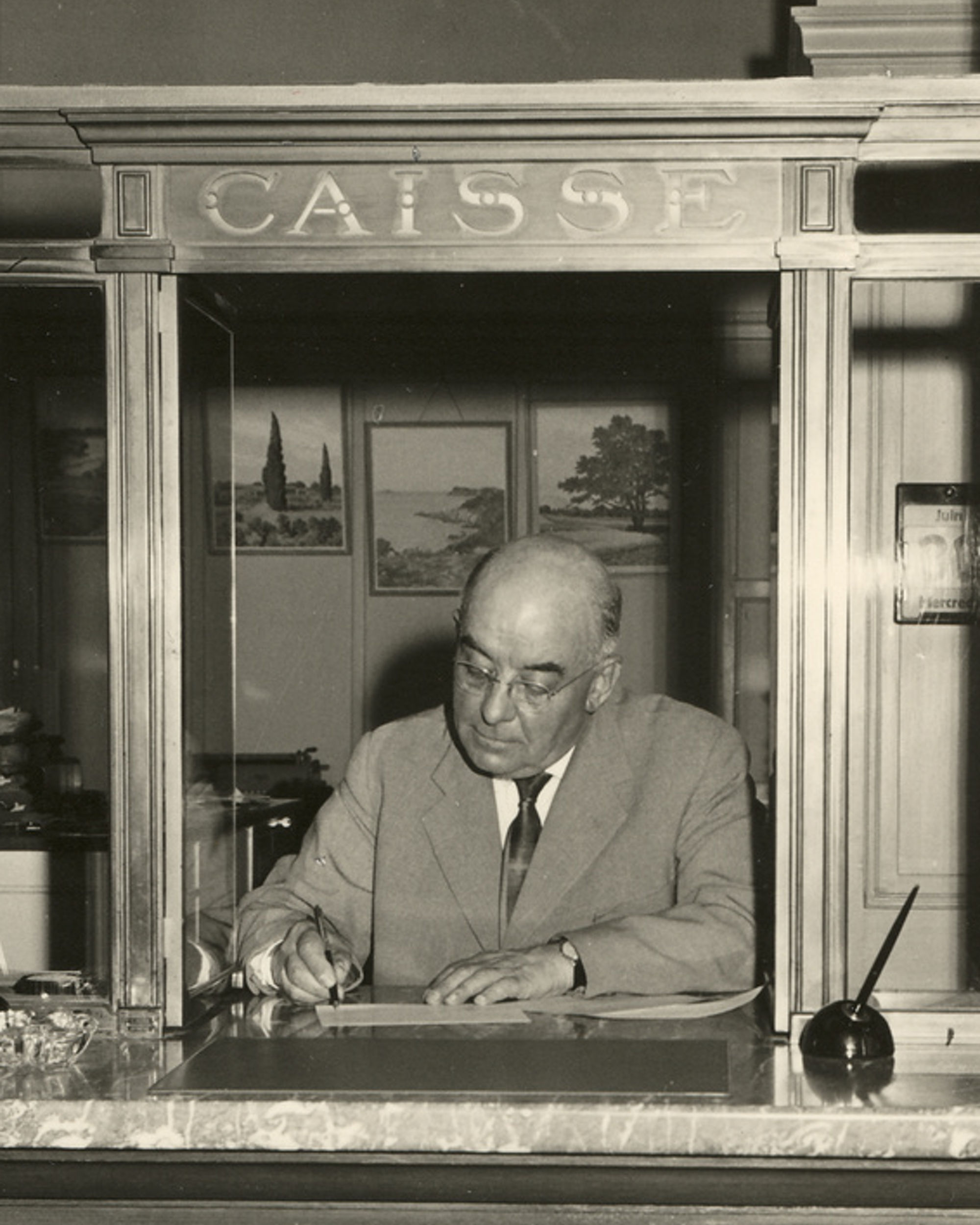Black and white photograph of a bank employee in a suit, behind a counter marked ‘Cashier’.