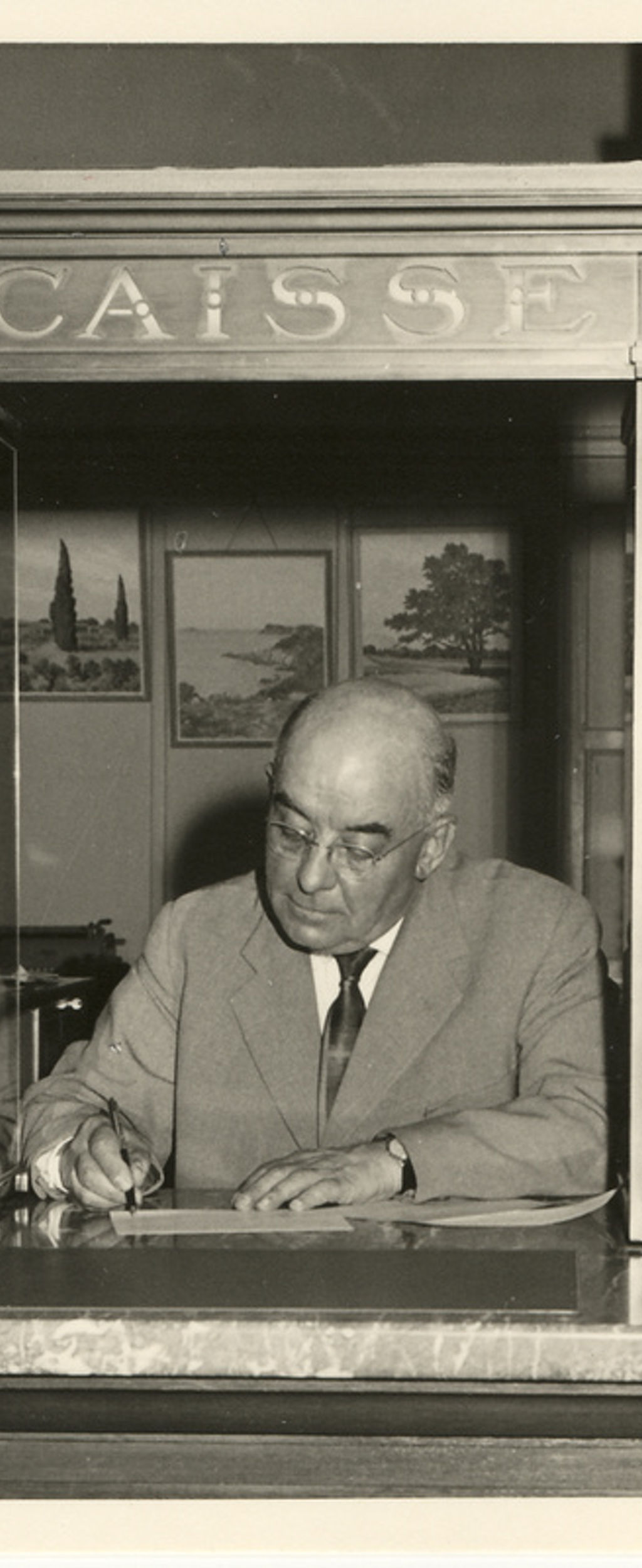 Photo en noir et blanc, un monsieur en costume, employé de banque, derrière un comptoir surmonté de l'inscription "Caisse"