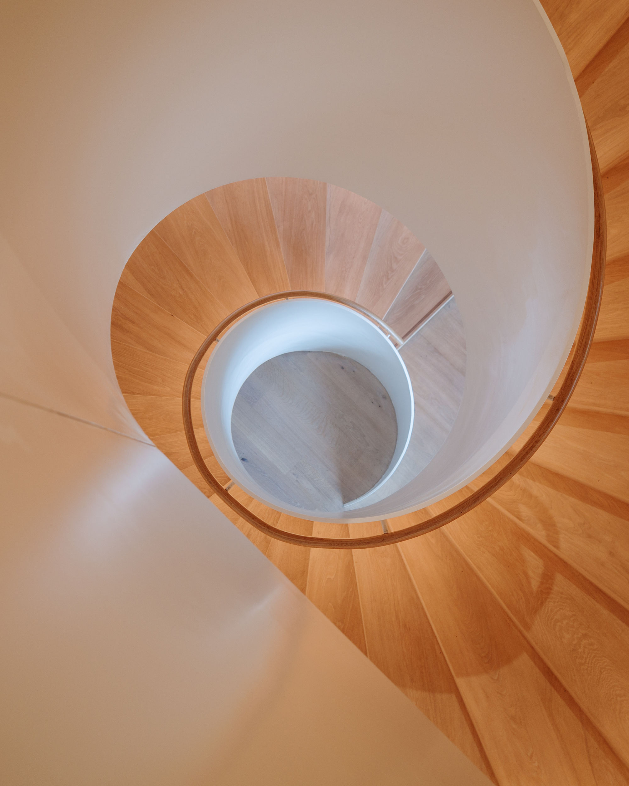 Vue plongeante sur un escalier en colimaçon au style moderne, composé d’une rampe pleine de couleur blanche et de marches en bois clair, dont l’enroulement crée un motif architectural hypnotique.